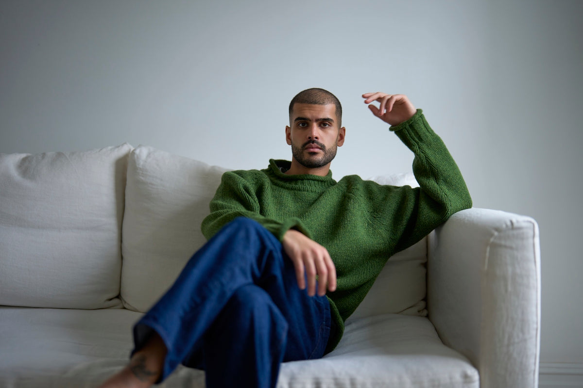 Man sitting on a white couch wearing a green sweater and blue pants.