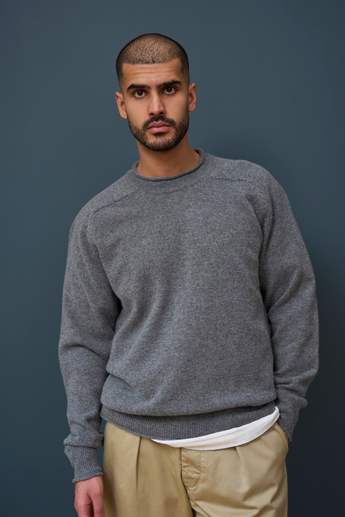Man wearing a gray sweater and beige trousers against a dark background
