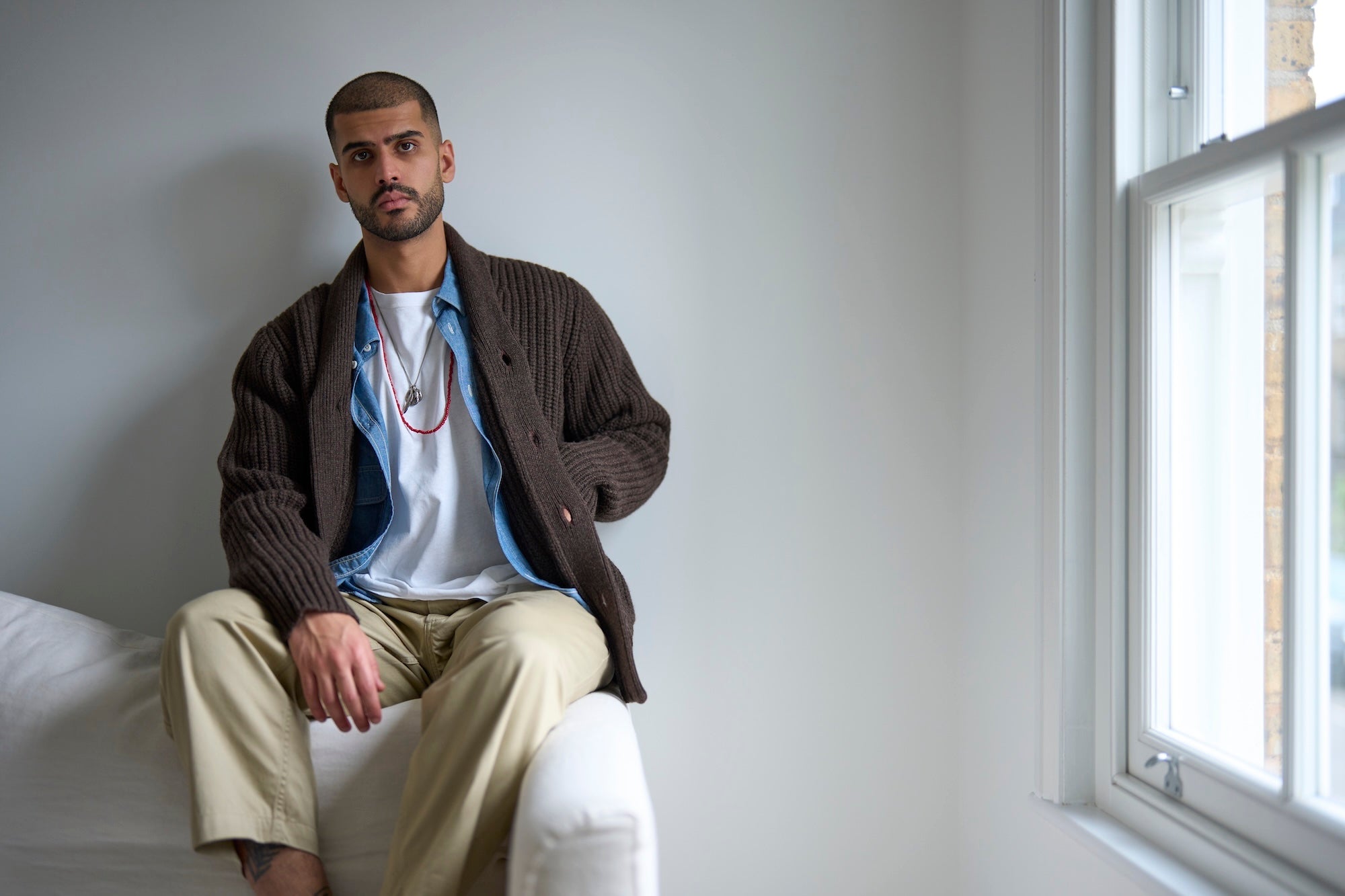 Man sitting on a white couch wearing a brown jacket and beige pants in a room with a window.