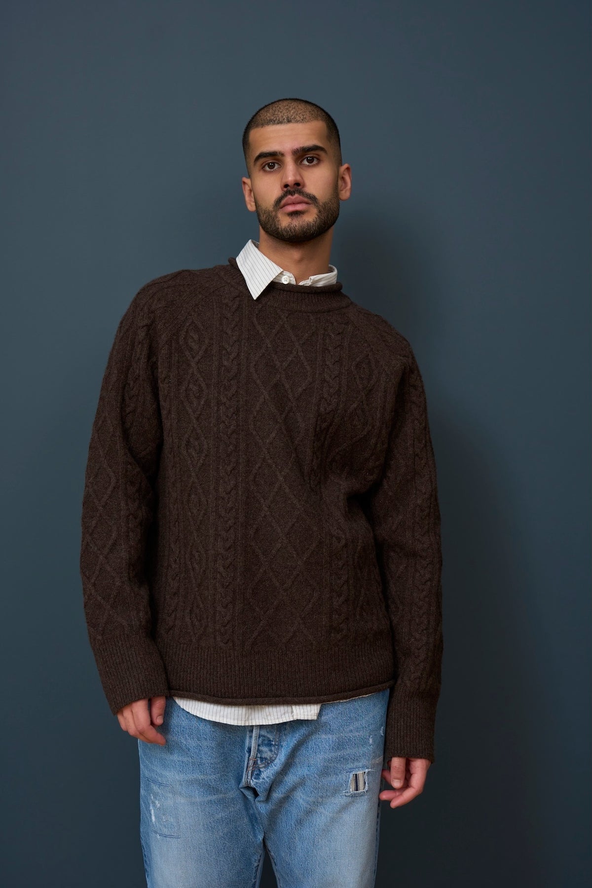 Man wearing a brown cable knit sweater over a white shirt with blue jeans against a gray background